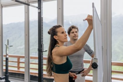 Female fitness coach writes on whiteboard as a male athlete watches in a gym.